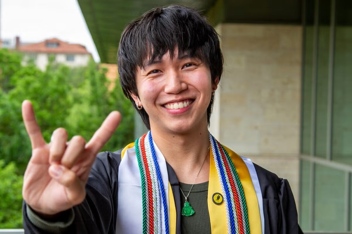 A student with a pin, multipel cords, and graduation gown and science stoll holds up the hook 'em hand symbol on the UT campus.