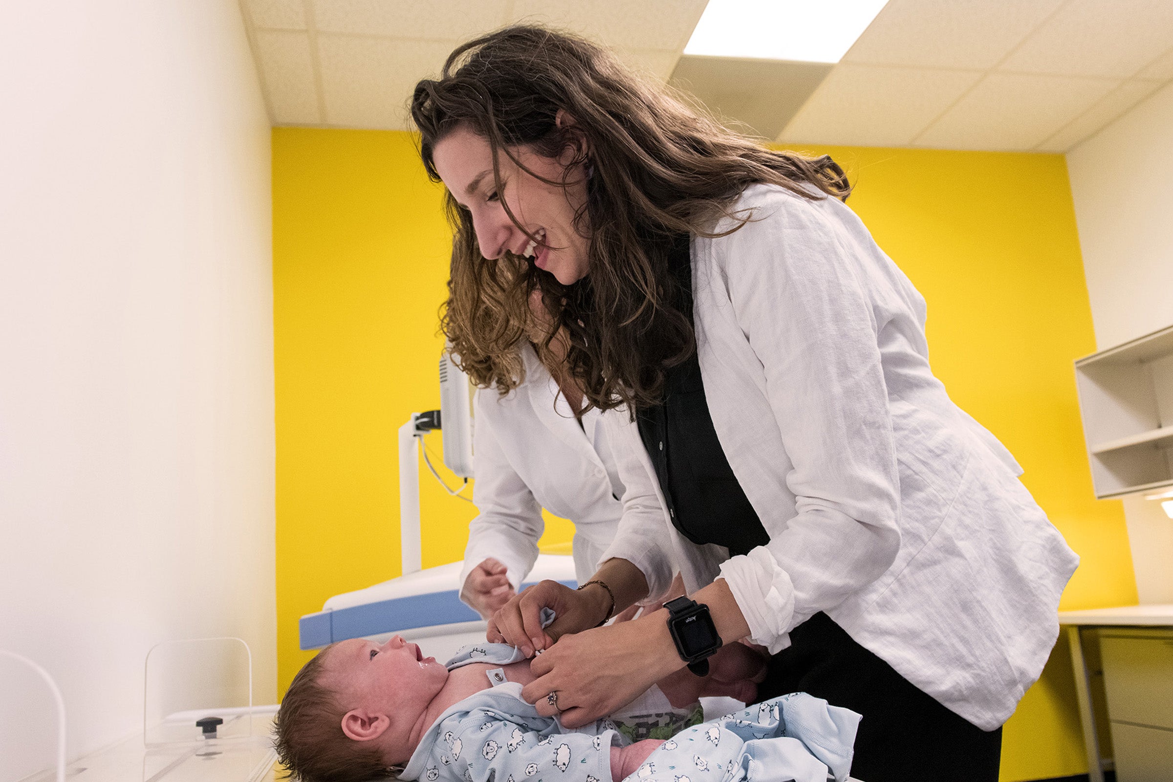 Beth Widen, a dark haired woman in a white coat, leans over an infant lying on a table. She smiles down at the infant.
